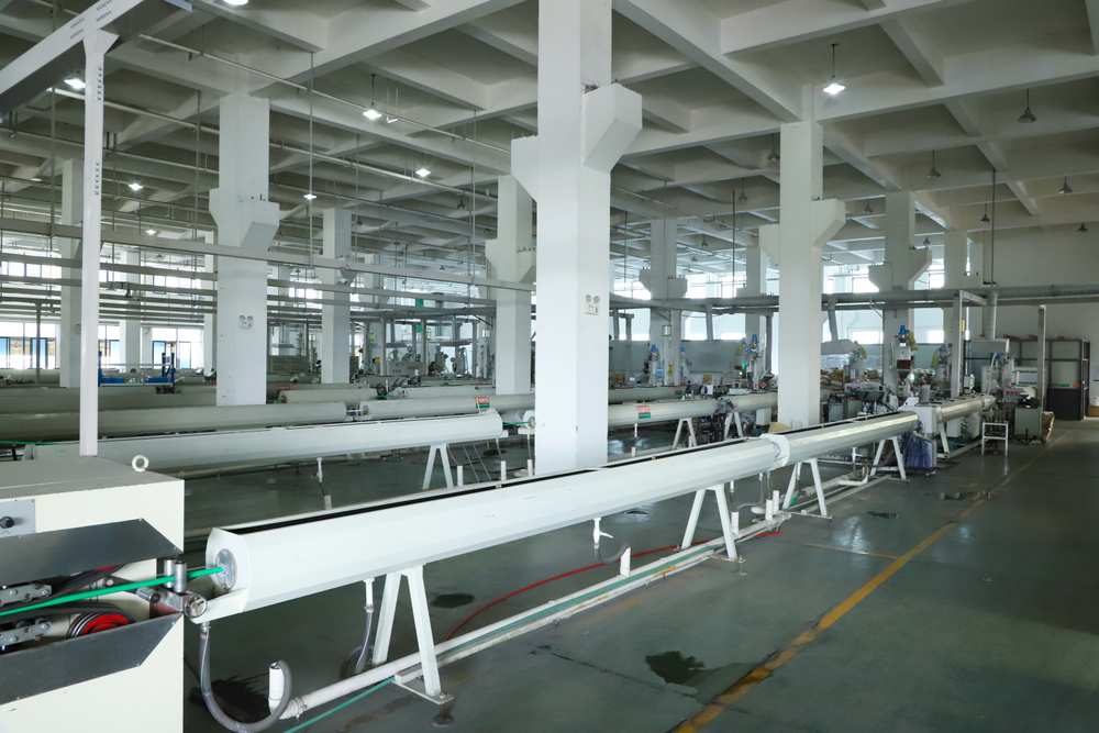 A white plastic pipe running along the support stands of an extrusion line in a long factory hall at IFAN&rsquo;s plant