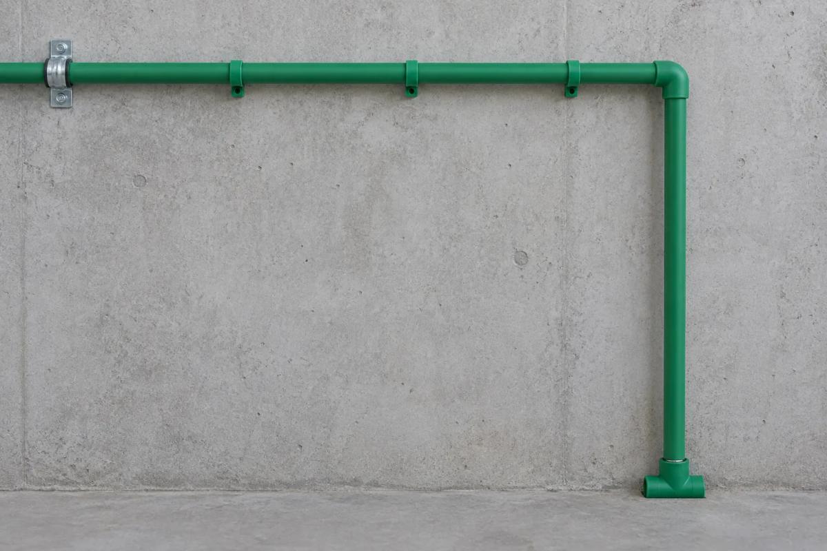 Illustration of an exposed green PP-R pipe on a concrete wall: a steel clamp at the far end, open guide clips along the run, and an elbow turning into a free vertical leg