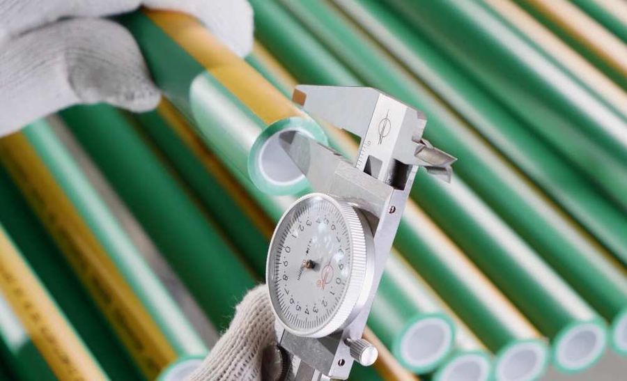 Gloved hands measuring the wall thickness of a green PP-R pipe with a dial caliper across a cut end, with mixed green and yellow-striped pipe stacked behind