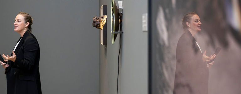 A curator stands with a microphone near a large work of art, photographed at an angle such that only light reflecting off the glass is visible. Other artworks are installed in the distance on dark grey walls.