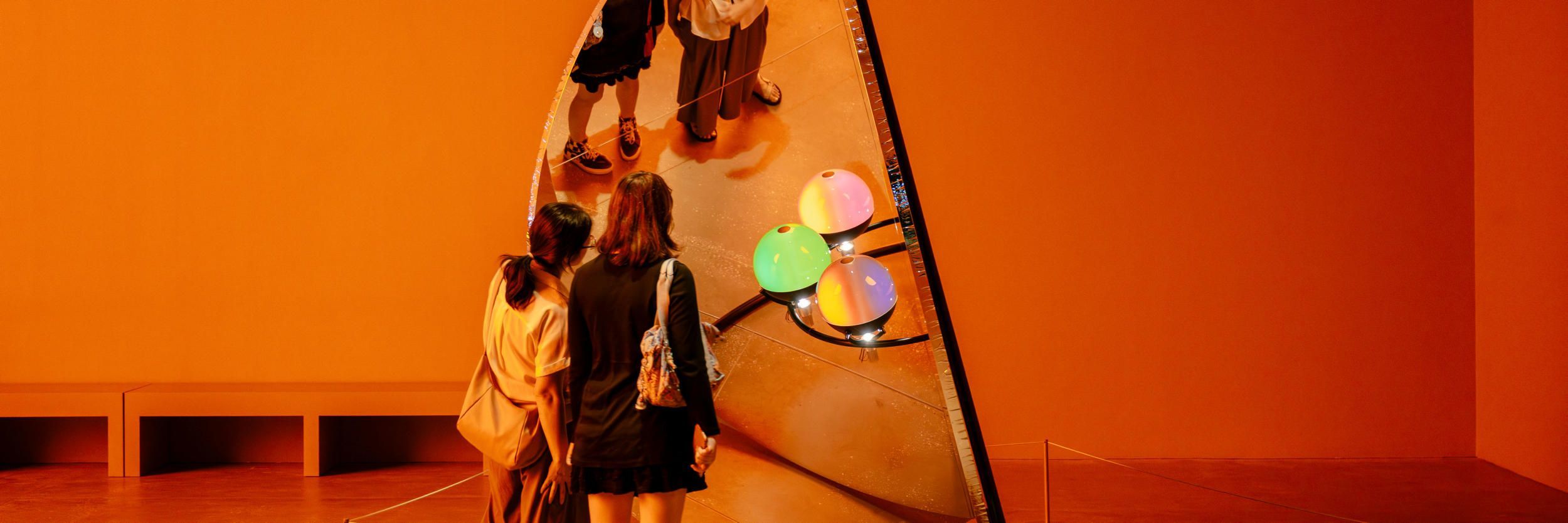 Two people view a circular frame holding glossy coloured spheres, which appear blue, teal, green and amber, while multiple reflections of their figure appear in the tall mirror behind the arrangement.