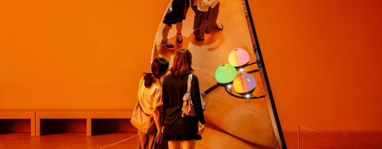 Two people view a circular frame holding glossy coloured spheres, which appear blue, teal, green and amber, while multiple reflections of their figure appear in the tall mirror behind the arrangement.