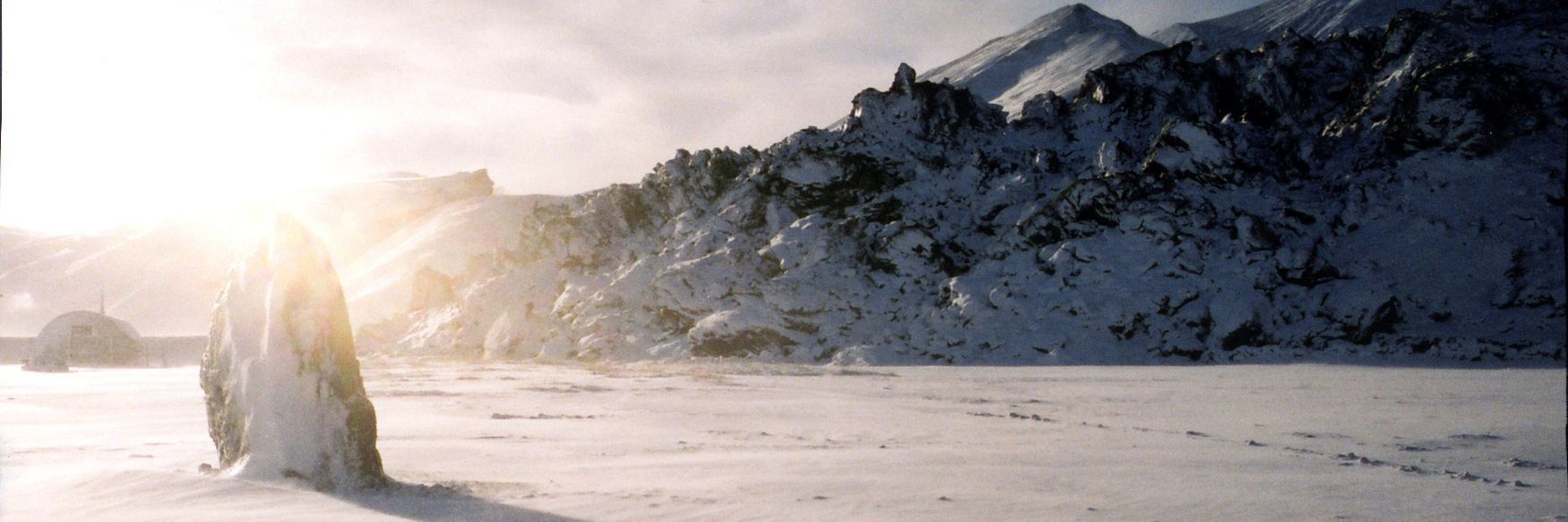 A tall, snow-covered rock stands in a wide, frozen landscape with a long shadow stretching across the snow, illuminated by low sunlight against a backdrop of rugged, snow-covered mountains and a dome-shaped structure.