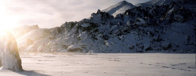 A tall, snow-covered rock stands in a wide, frozen landscape with a long shadow stretching across the snow, illuminated by low sunlight against a backdrop of rugged, snow-covered mountains and a dome-shaped structure.