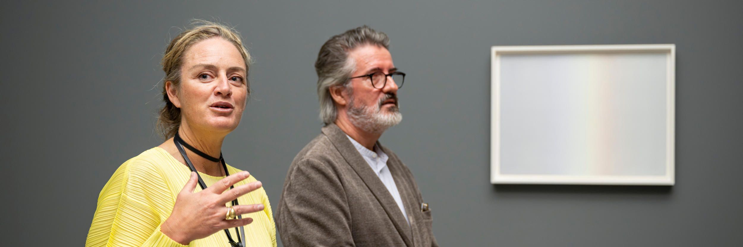 Two people stand beside artwork, installed on a grey wall, with a vertical gradient with faint rainbow colours gradually progressing from cool grey-blue to warm cream.