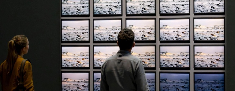 Two people view a series of 20 photographs installed in rows of five on a dark gallery wall; the photographs depict snowy landscapes in Iceland.
