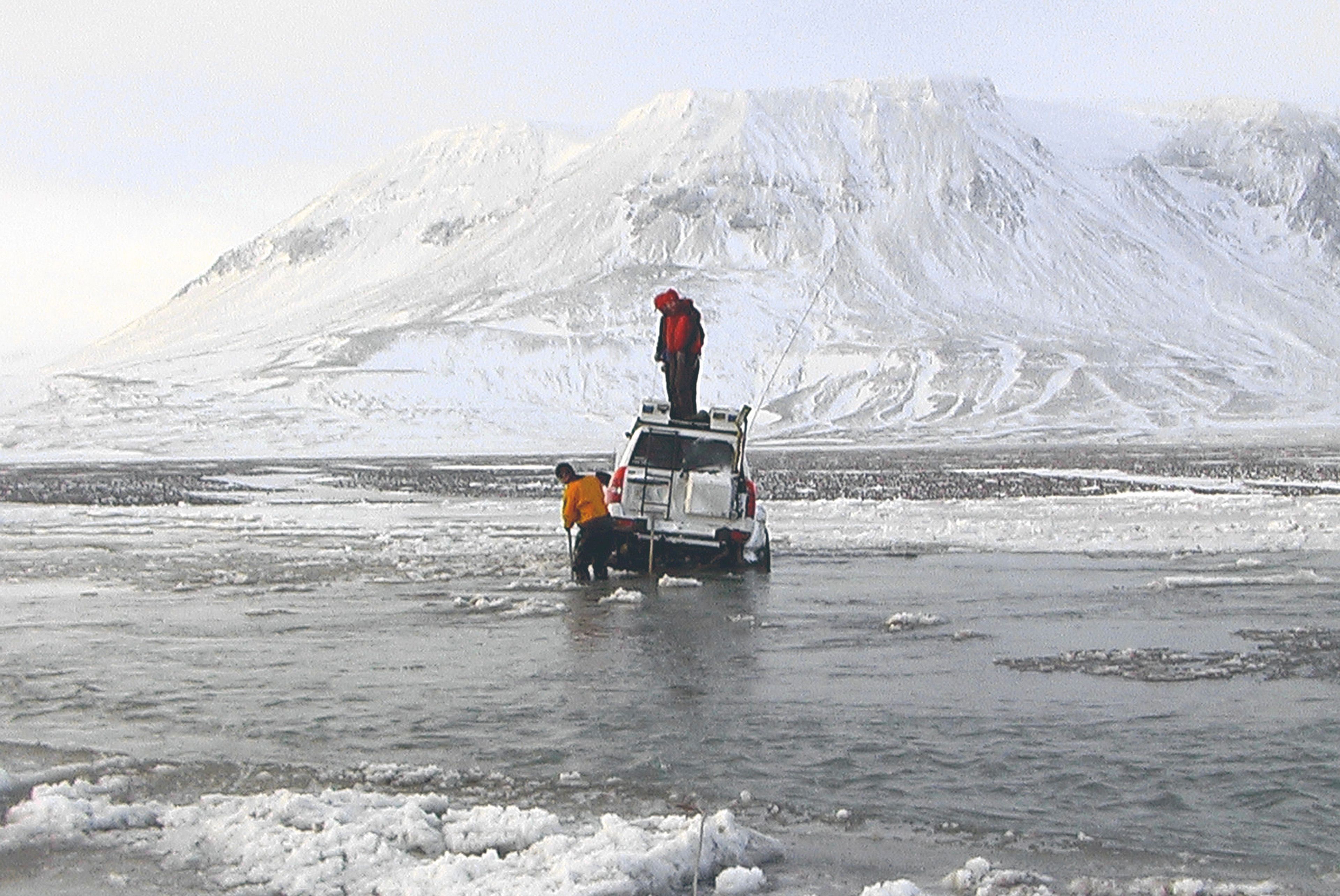 A person in a hooded rain jacket stands on the roof of a white off-road vehicle, which is tilted and sunk in icy water, while another person in an orange jacket stands knee-deep in water to the side. Snowy mountains rise in the distance.