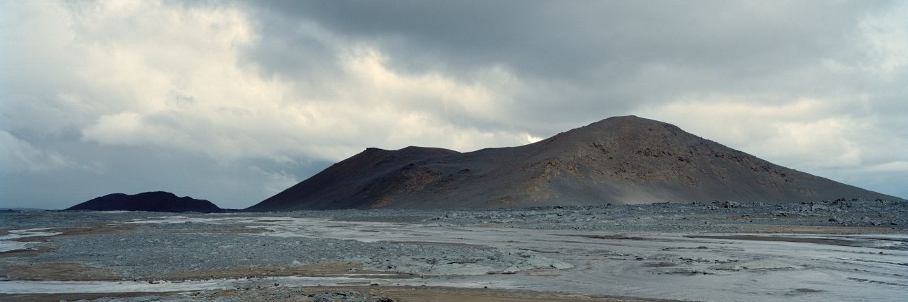 A wide, muddy wetland plain with branching, water-filled channels weave toward distant low hills, with a cloudy sky above.