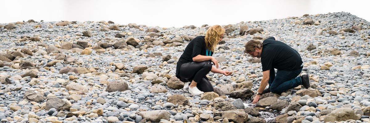 A person crouches on a rocky surface and extends a hand into a thin stream of running water that winds between grey and tan stones in a white-walled gallery.