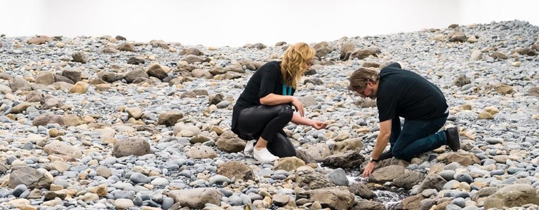 A person crouches on a rocky surface and extends a hand into a thin stream of running water that winds between grey and tan stones in a white-walled gallery.