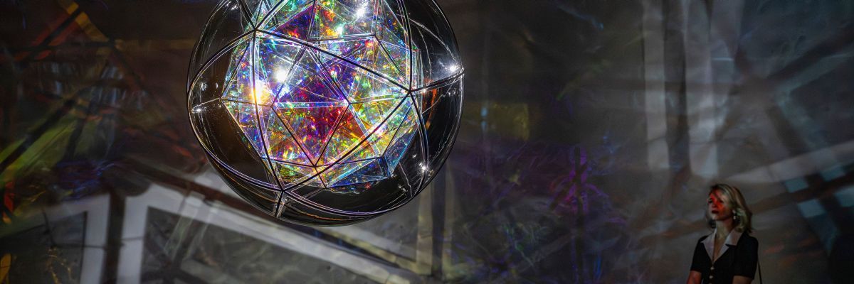 The multi-faceted glass form shimmers with intense light, each pane reflecting different saturated colours within the mirrored interior, creating a dense field of rainbow colours. A visitor looks on from the right-hand side.