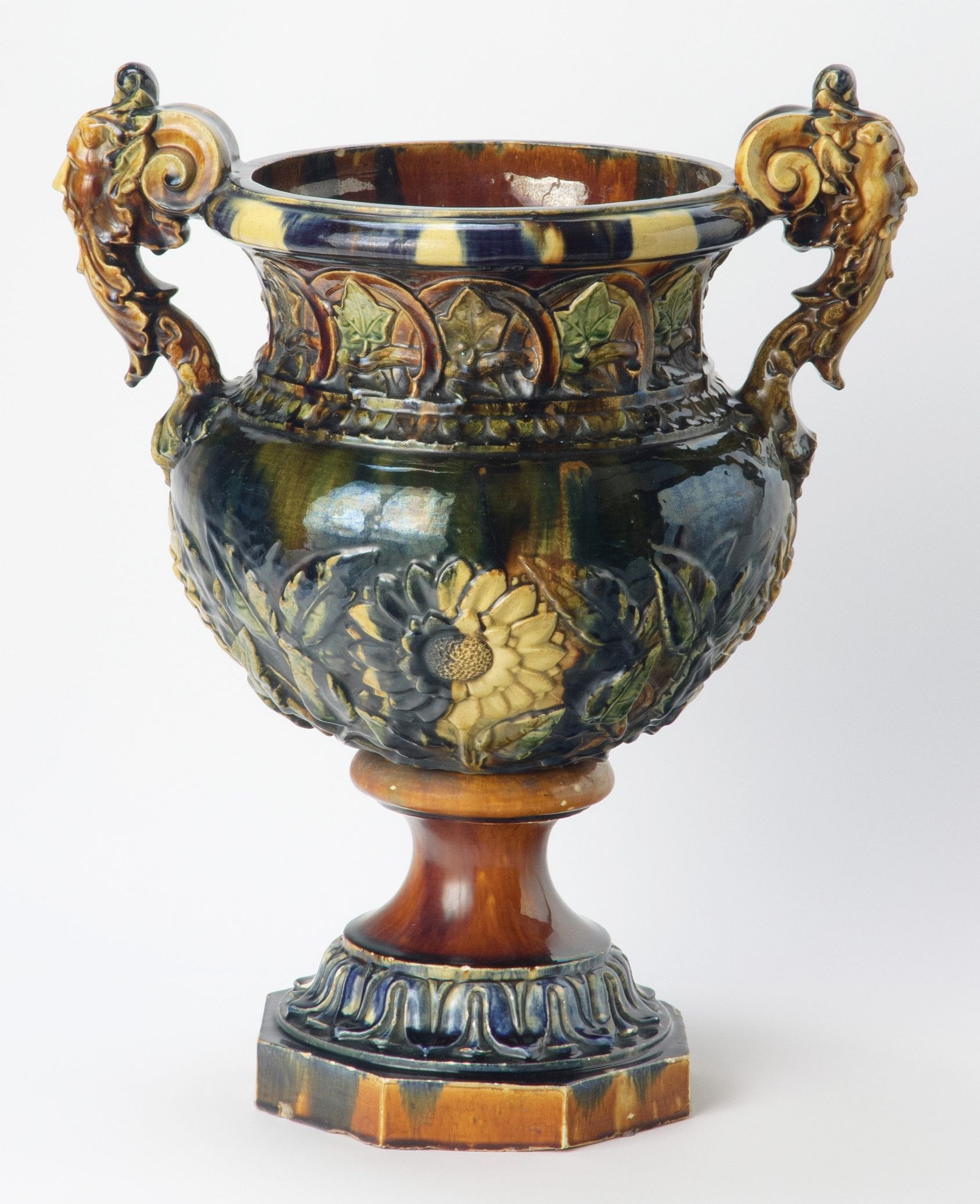 JL (James Laurence) Watts, Queensland Australia 1849–1925 / James Campbell & Sons, Queensland Australia 1885–1948 / Jardiniere c.1890 / Earthenware white clay body. The neck modelled with ivy leaves within arches, the swelling body with four sunflowers and leafy branches and the handles modelled as two grotesque masks. Glaze splashed clear brown, green and deep cobalt / 61 x 49 x 32.2cm / Gift of Miss Alison Campbell 1986 / Collection: Queensland Art Gallery | Gallery of Modern Art