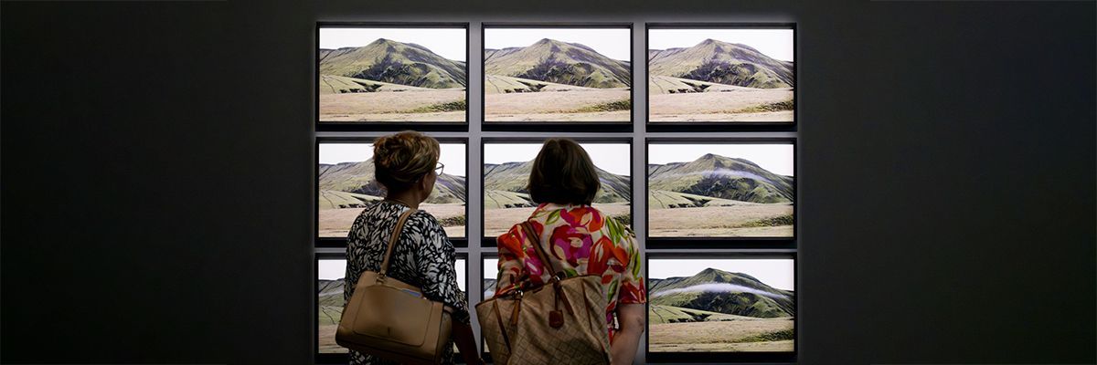 Two visitors view a grid of nine framed photographs, arranged in a grid of three rows by three columns, showing sequential photographs of a green mountain as a cloud forms and passes across its slopes.