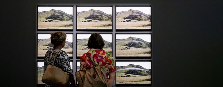 Two visitors view a grid of nine framed photographs, arranged in a grid of three rows by three columns, showing sequential photographs of a green mountain as a cloud forms and passes across its slopes.