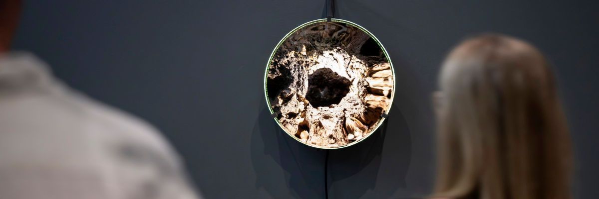 A person views a dark, porous stone, which hangs from a thin black cord in front of a large concave mirrored disc, whose rippled metallic surface reflects distorted light and crater-like textures, with the disc held by small metal clips and suspended from a vertical metal bracket on a dark grey wall.