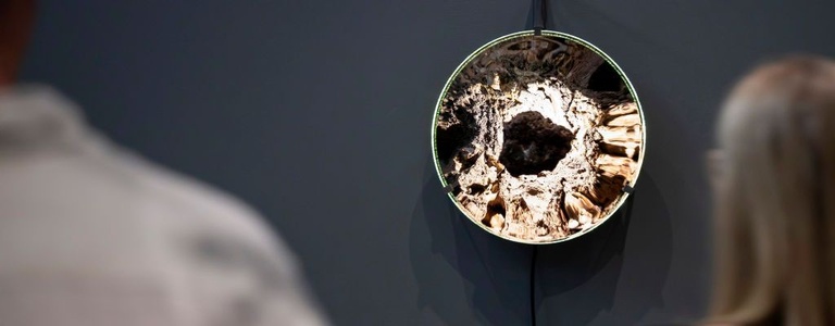 A person views a dark, porous stone, which hangs from a thin black cord in front of a large concave mirrored disc, whose rippled metallic surface reflects distorted light and crater-like textures, with the disc held by small metal clips and suspended from a vertical metal bracket on a dark grey wall.