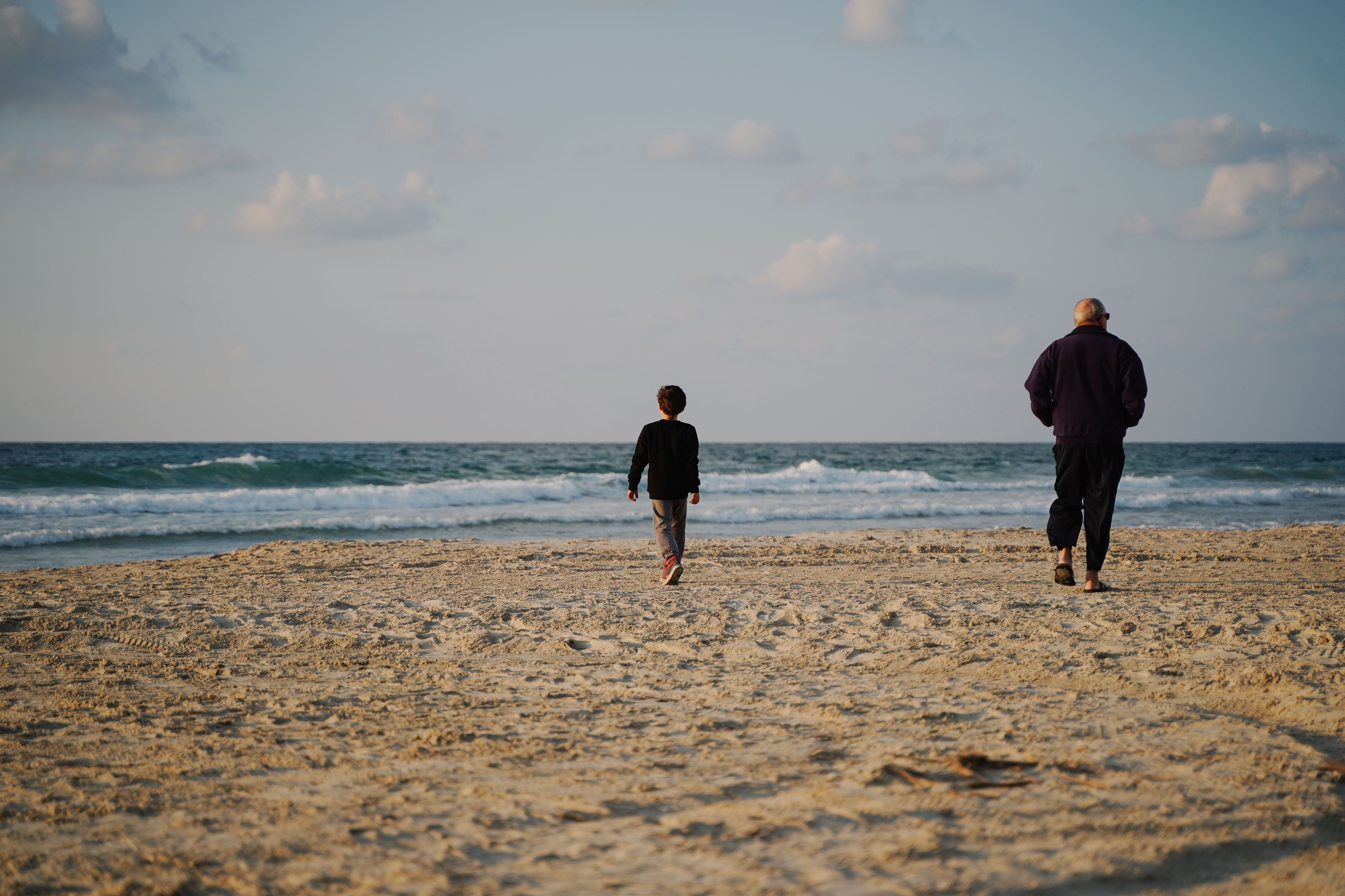 Image An older man and a young boy walk side by side toward the ocean on a sandy beach at dusk, viewed from behind.
