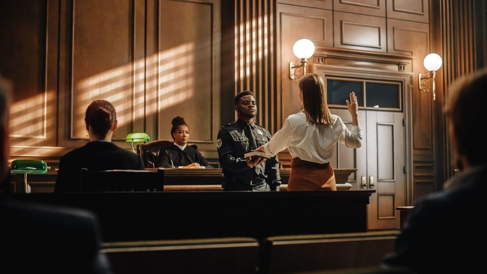 A witness raising their hand to testify in a courtroom as a judge and officer observe during a domestic violence case.