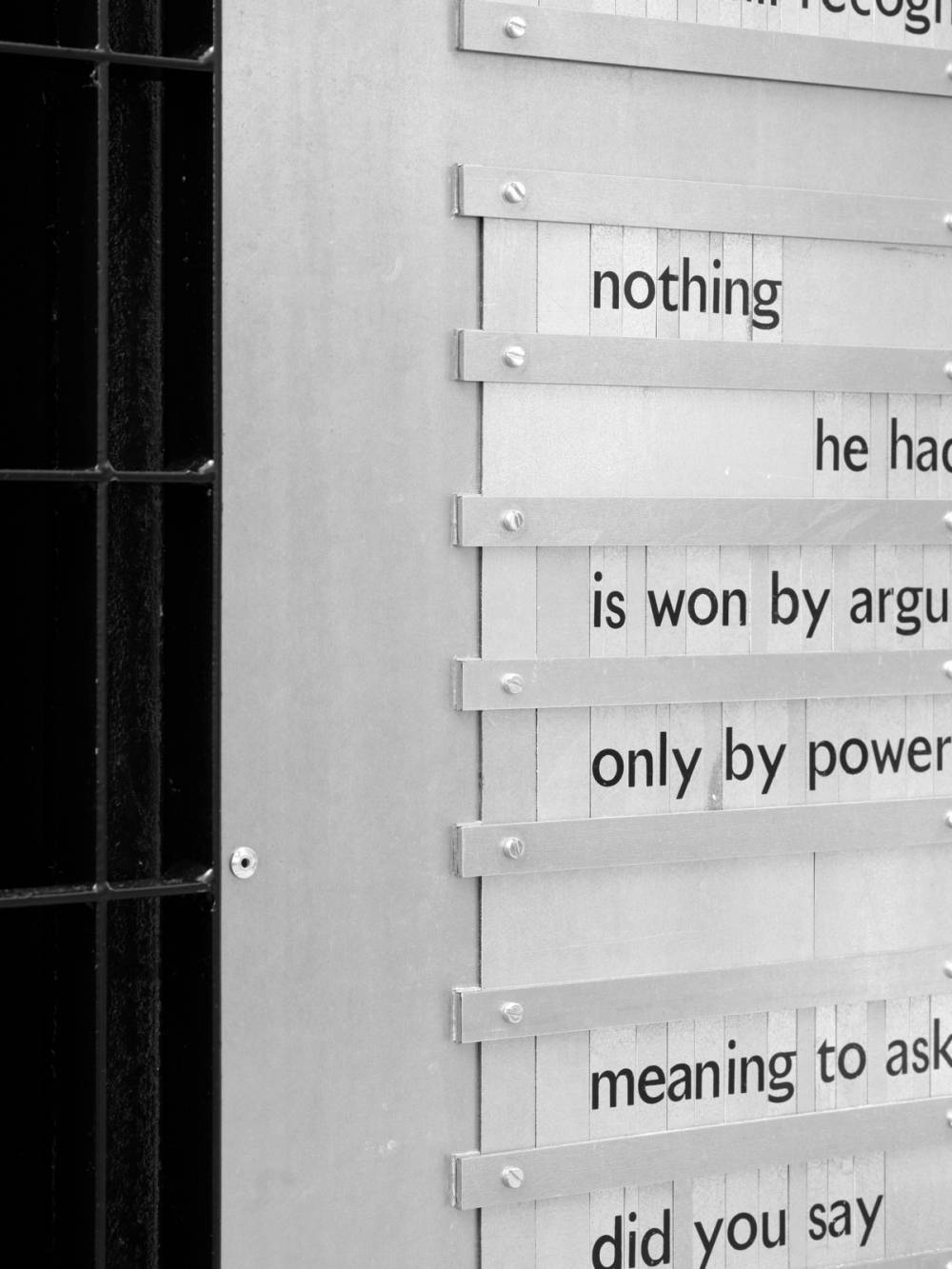 A monochrome detail of a spatial installation, juxtaposing a dark metal grate on the left with a lighter, textured assembly. This assembly features a smooth metal panel bordering vertical strips, which are secured by horizontal metal bars with exposed screws, creating a distinct layered form. Fragmented text is printed on the vertical strips.