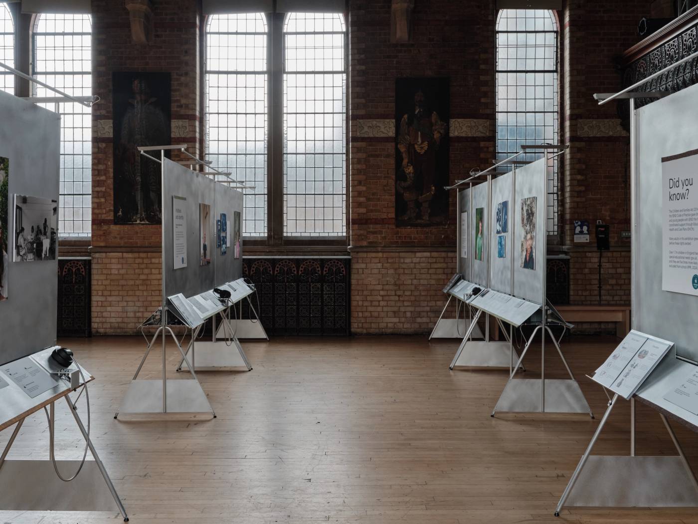 A photograph looking down a row of aluminium showcase structures displaying printed images and text panels. The background shows the ornate brick wall of a grand hall with large panelled windows.