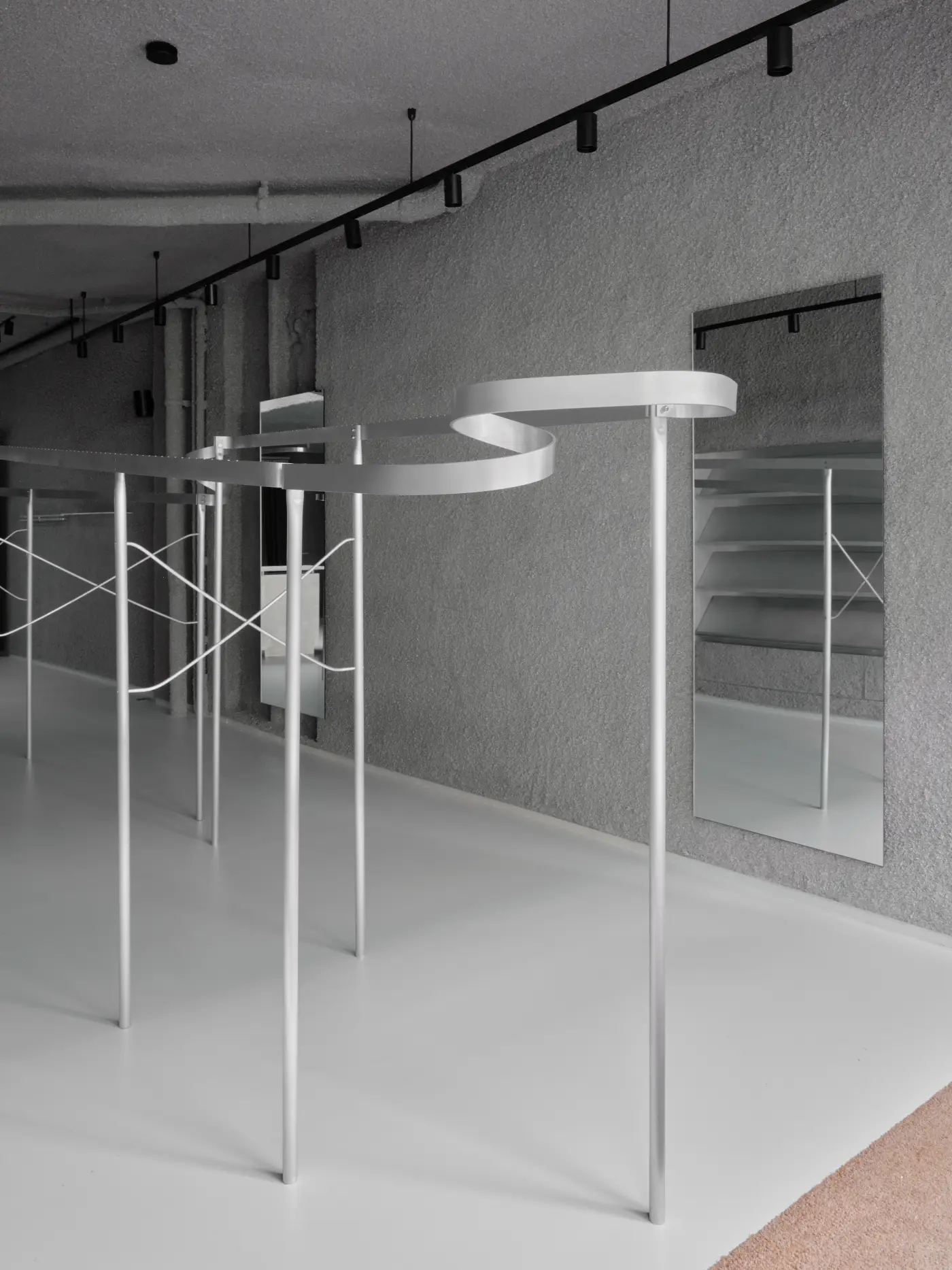 Minimalist menswear store interior featuring bespoke curving aluminum display rails and vertical poles on a light grey floor, contrasted by a textured dark grey sprayed paper pulp wall and black track lighting.