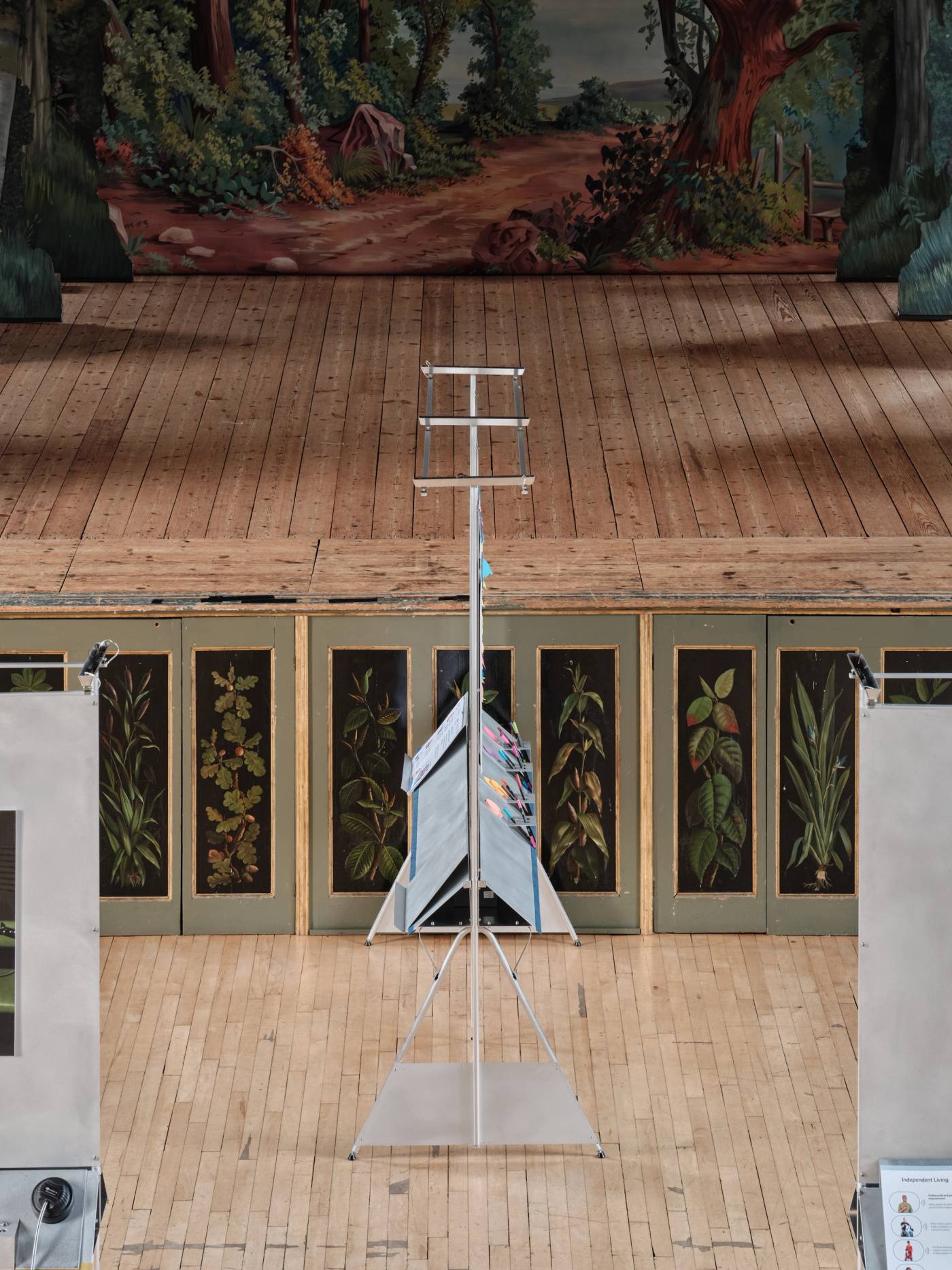 A photograph from an elevated position of an aluminium showcase structure from the side, displaying its triangular supports. The structure sits in front of a stage with wooden floorboards and ornate painted edge.