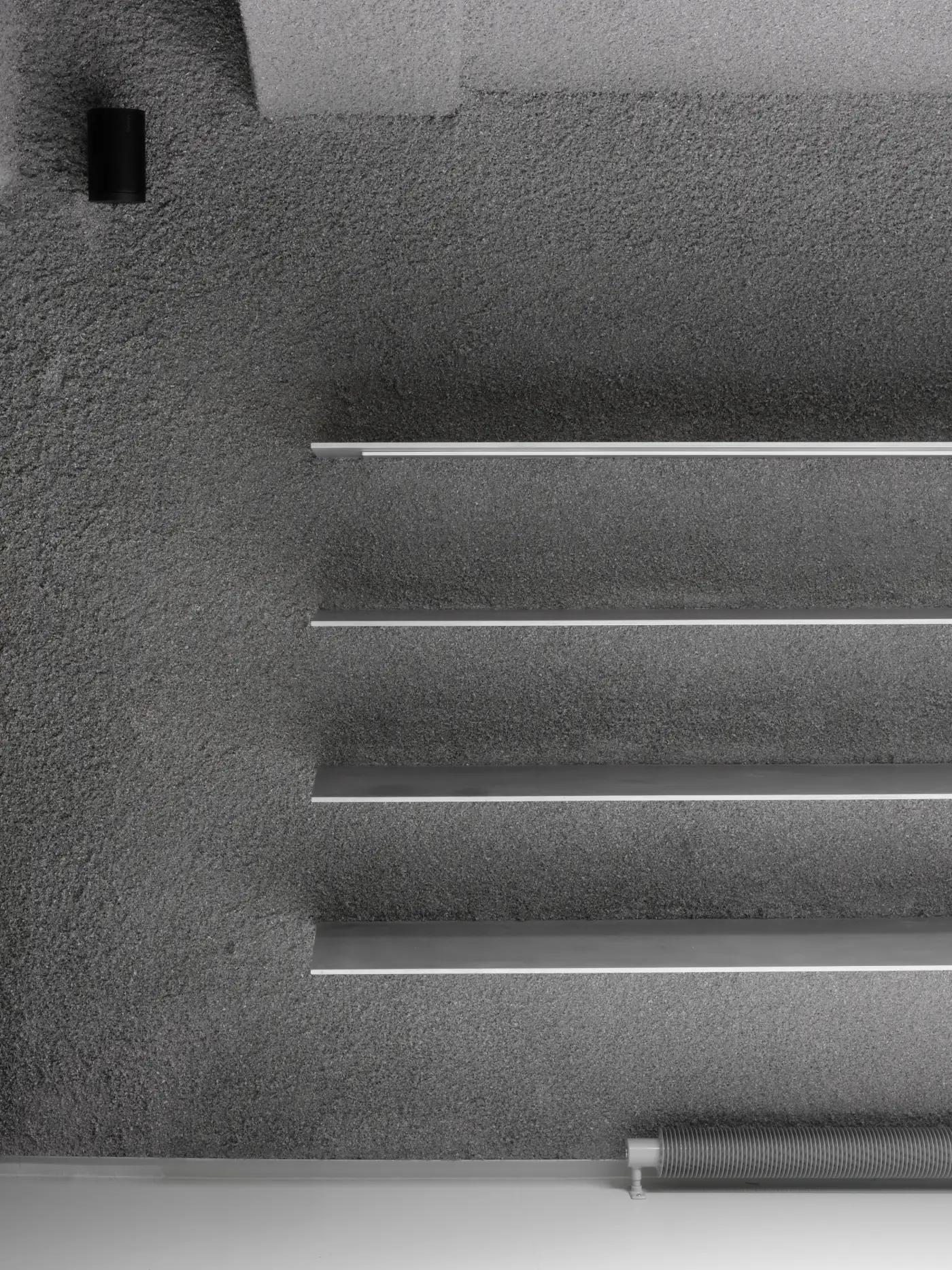 A richly textured, dark grey sprayed paper pulp wall features several sleek, horizontal aluminium rails, each with a subtle white strip along its upper edge, creating a refined display for a menswear store.
