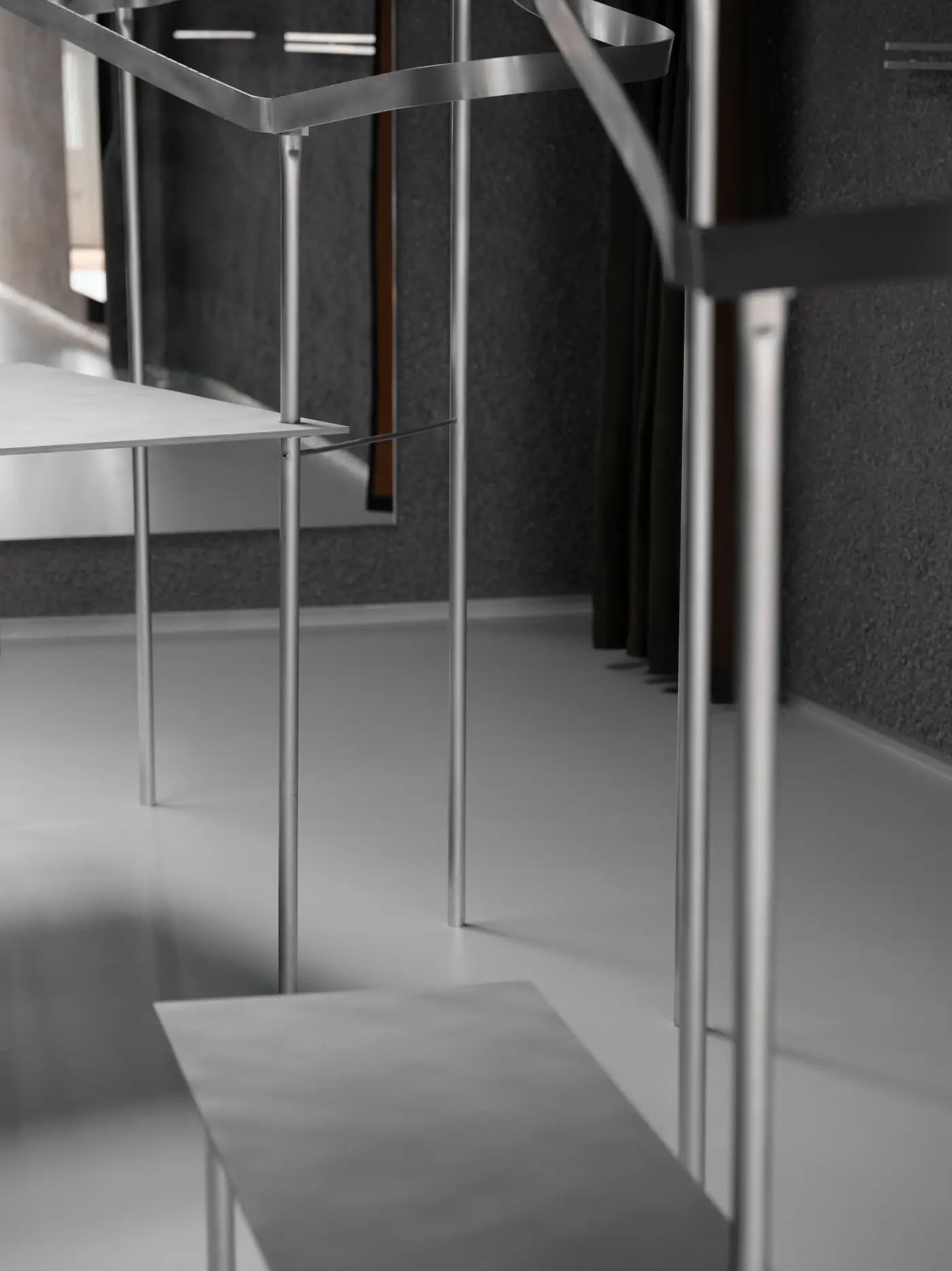 Bespoke slender aluminium rails and minimalist shelving units stand against a textured dark grey sprayed paper pulp wall and a smooth white floor in the Natalino menswear store.