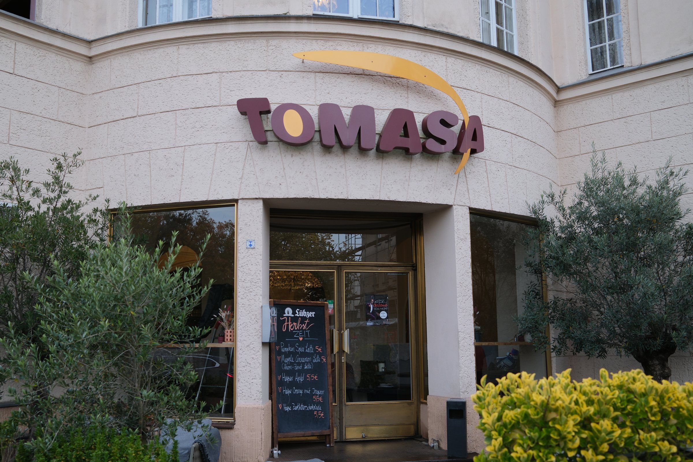 Eingang des TOMASA Zehlendorf mit Schriftzug, Olivenbäumen und Tafel mit Herbstangebot