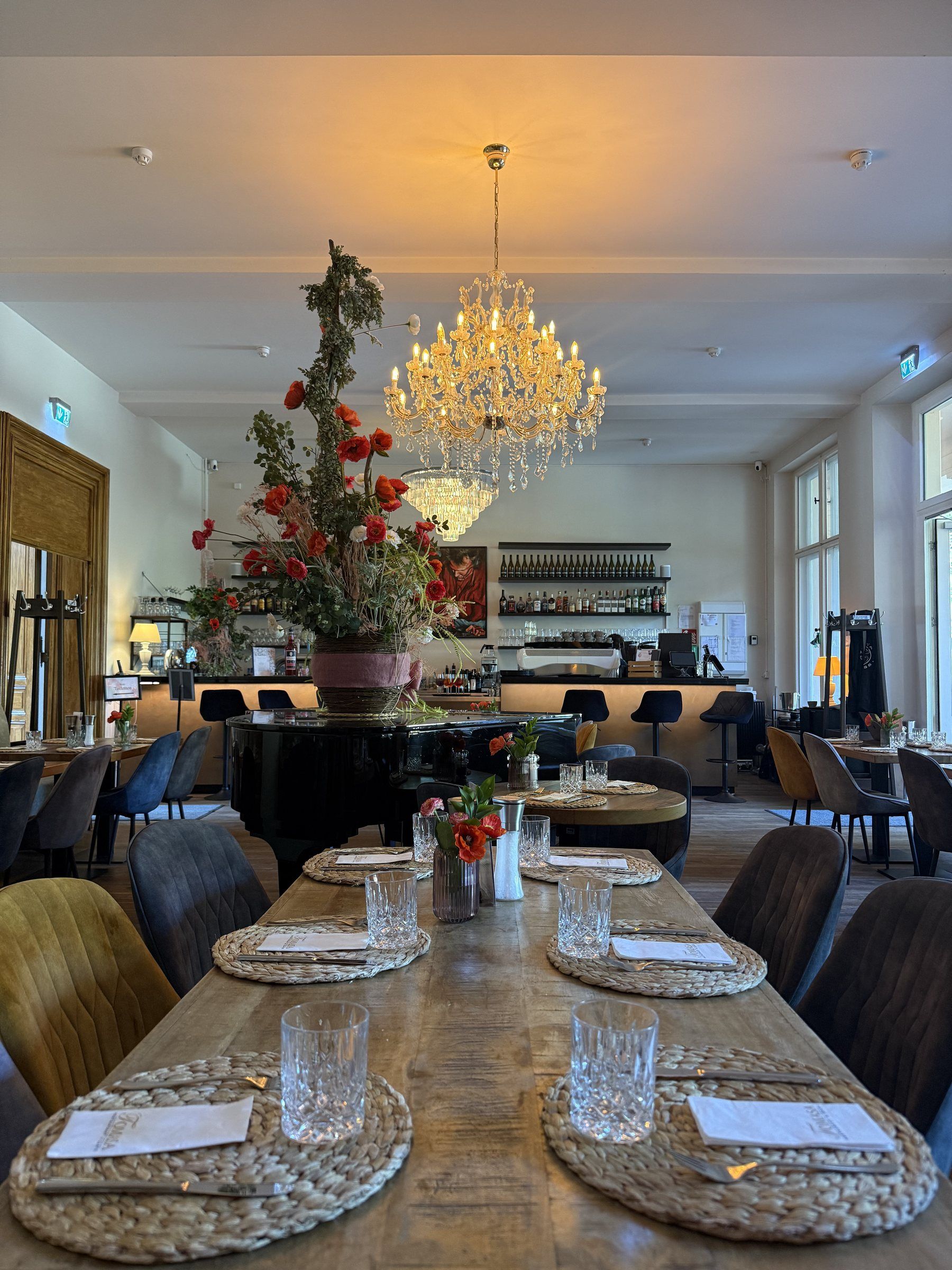 Der vordere Bereich mit langer gedeckter Tafel unter dem Kronleuchter, dahinter Flügel mit Blumenarrangement und die Bar