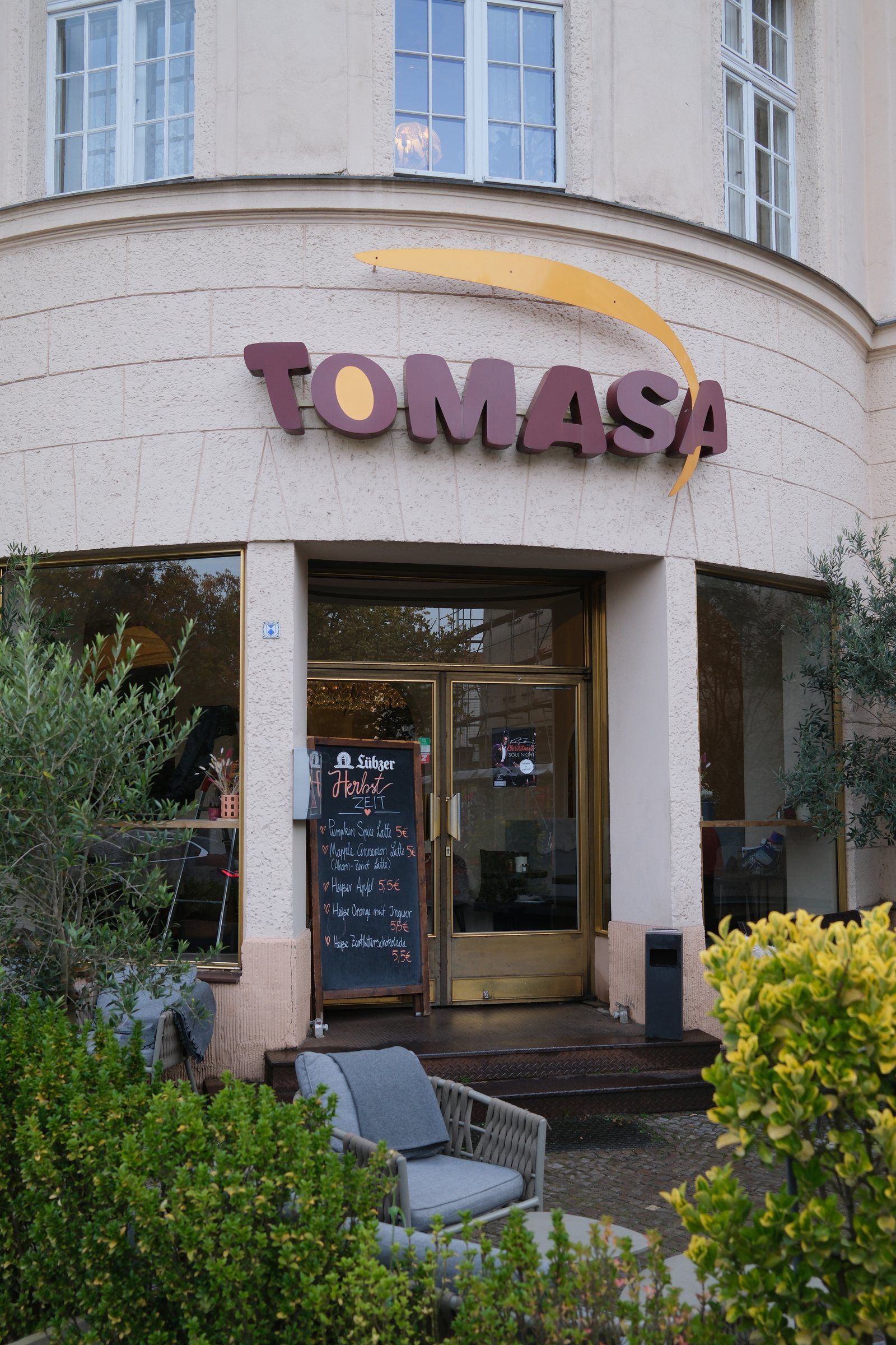 Eingang des TOMASA Zehlendorf mit Schriftzug, Olivenbäumen und Terrasse
