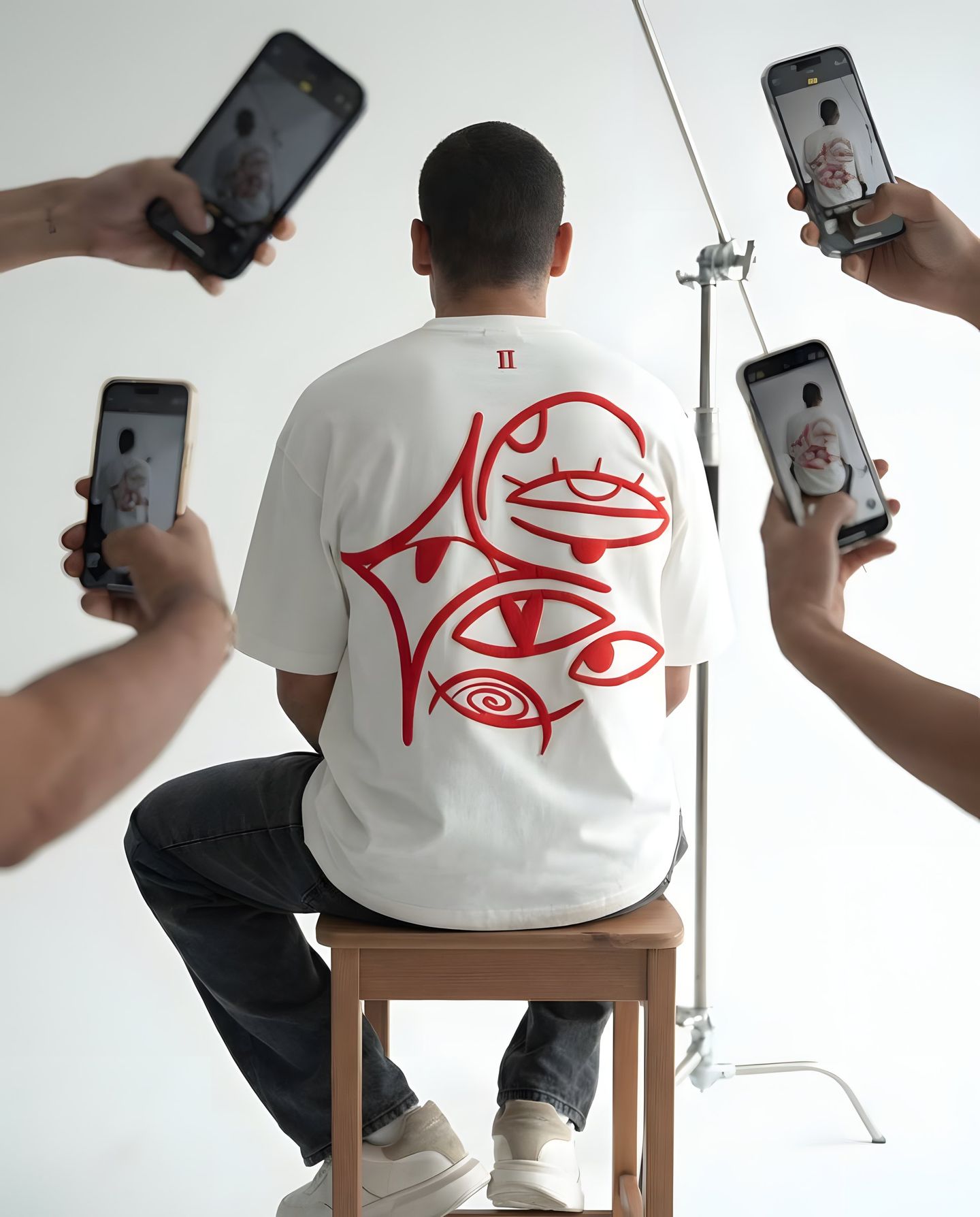 Seated model in a white Ariiston tee with red eye embroidery, filmed by five phones