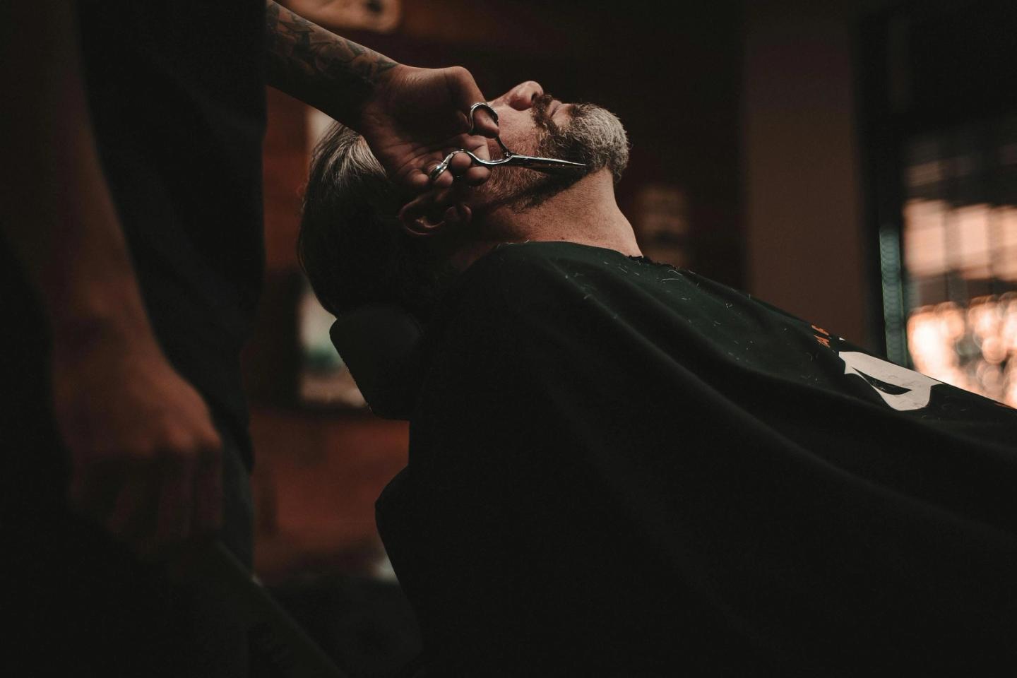A barber trimming a reclining client’s beard with scissors, in warm low light