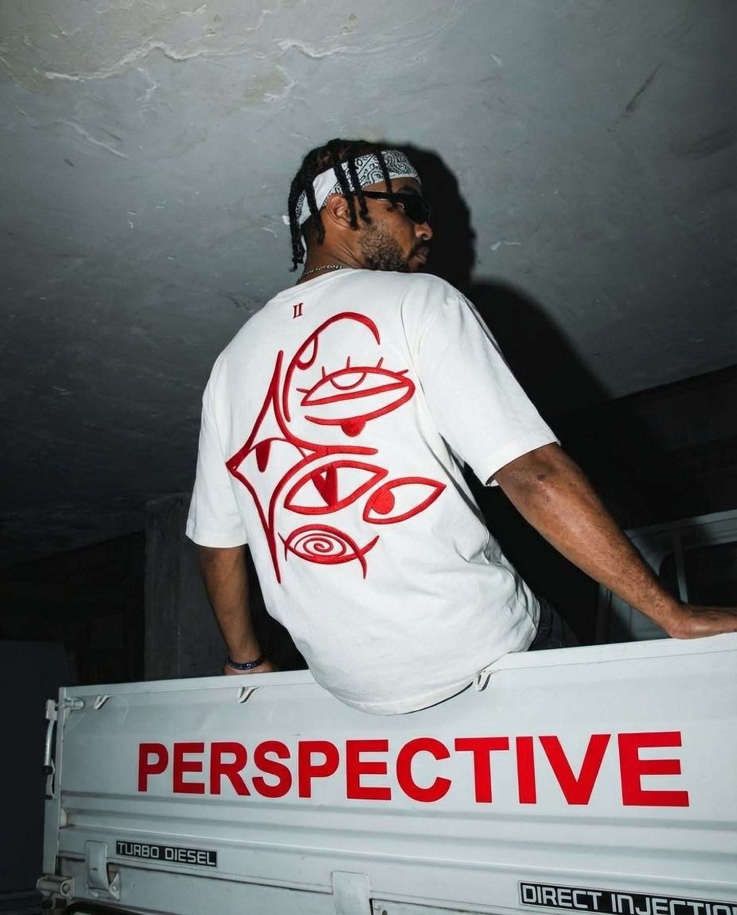 Model sitting on a truck tailgate that reads Perspective, in the white Ariiston tee with red eye embroidery