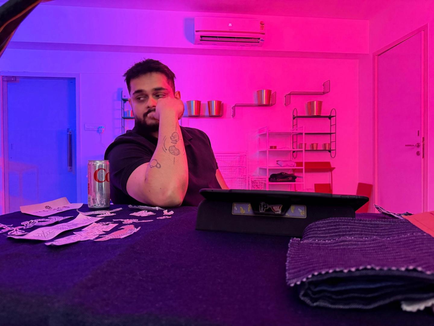 Kaustav at a desk under pink neon light, chin on his fist, a tablet and swatches in front of him