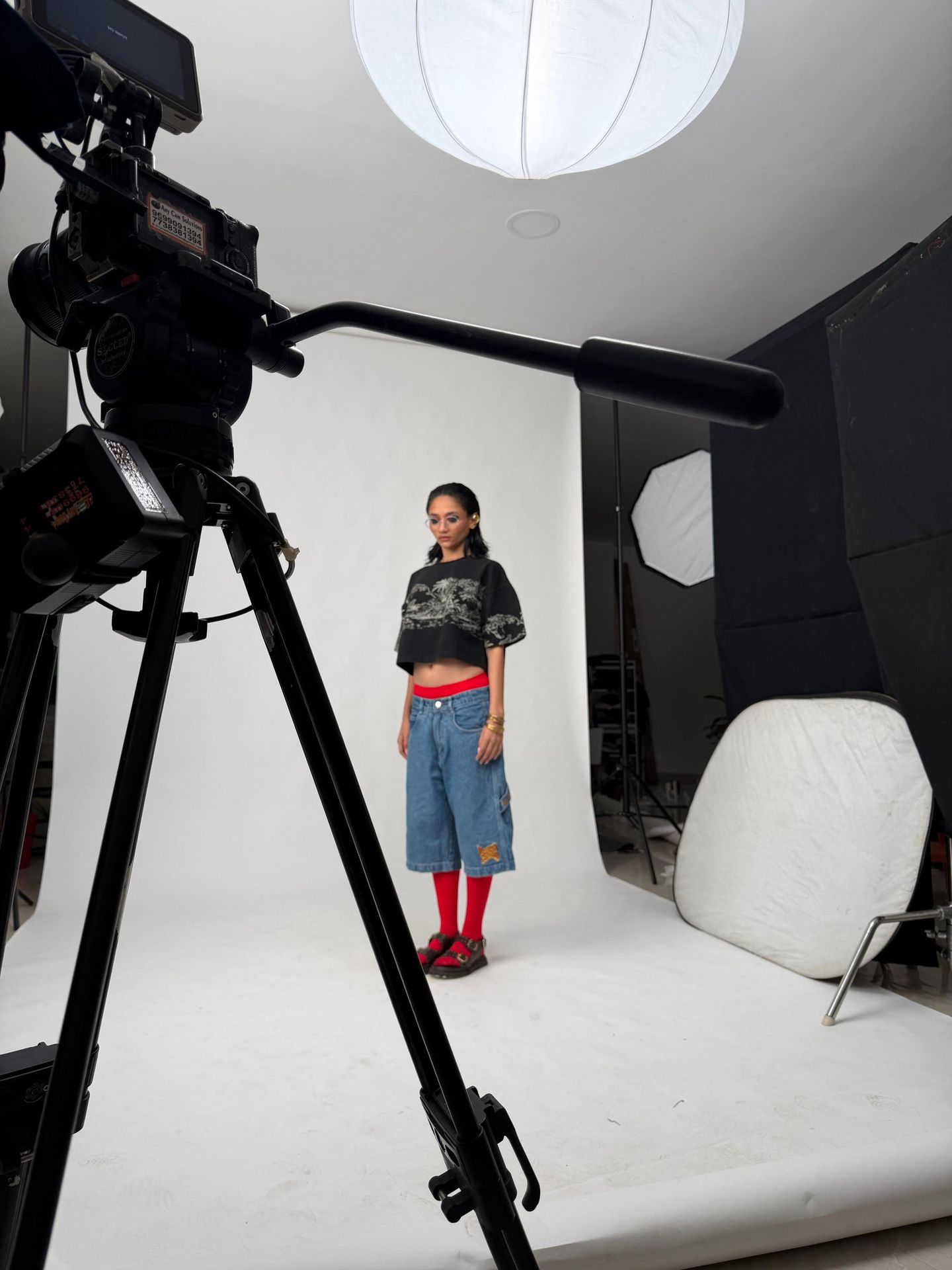 A model on a white studio backdrop, seen past the camera rig and lights