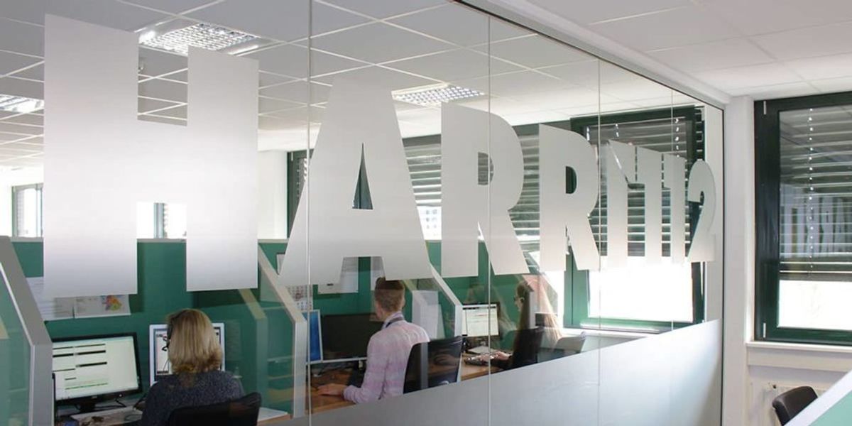 Angestellte sitzen hinter einer Glasscheibe, auf der das Harri-Logo groß aufgebracht ist.