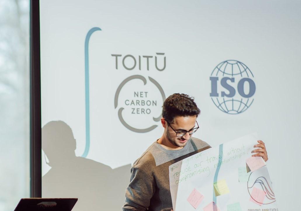 Photo of man with glasses holding sheet of paper standing in front of background showing Toitū's Net Carbon Zero mark and ISO logo