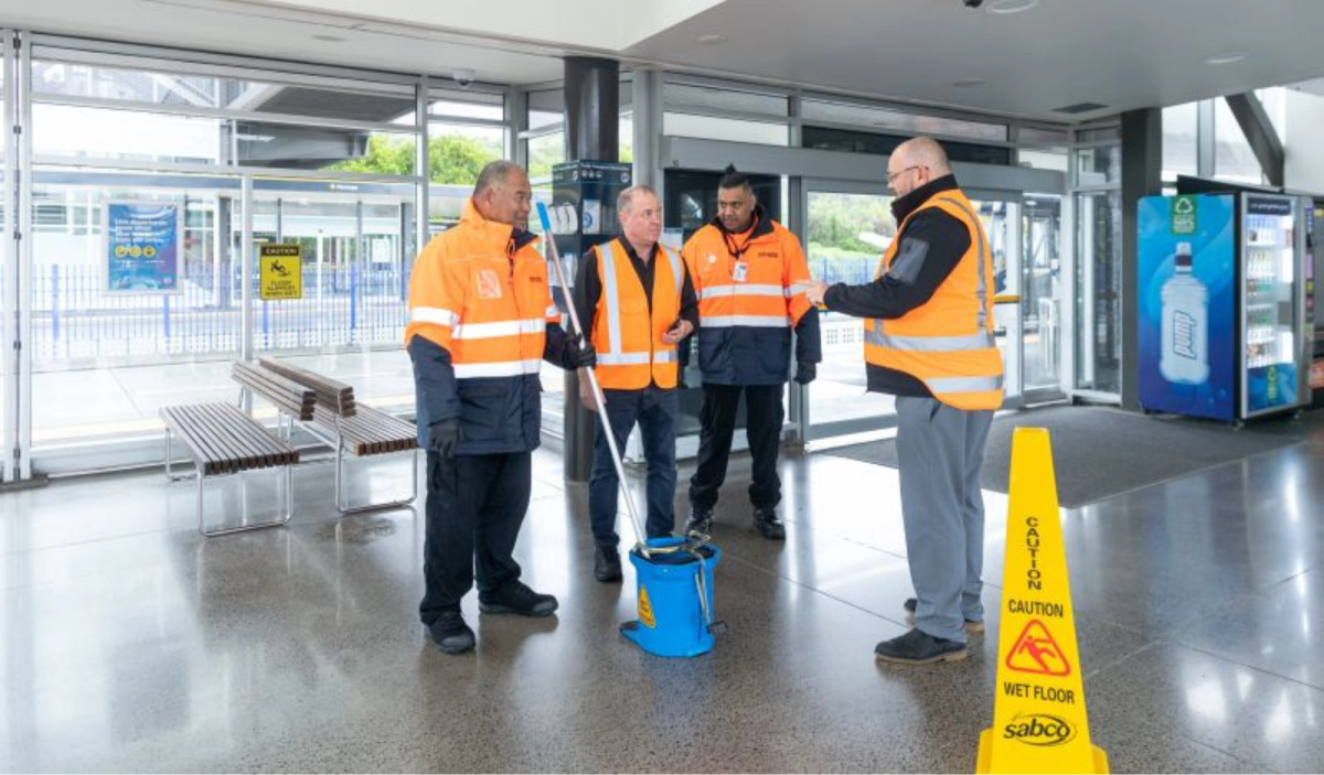 Four men in high vis jackets with mop and bucket and a caution wet floor sign on the floor in a bus station Four men in high vis jackets with mop and bucket and a caution wet floor sign on the floor in a bus station