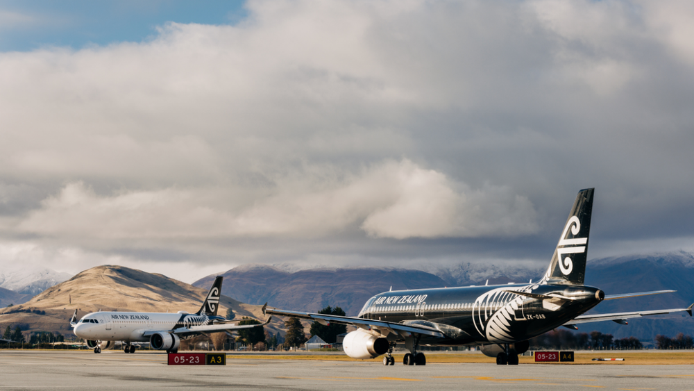 Two Air New Zealand planes sit on the runway tarmac in front of a mountainous New Zealand landscape