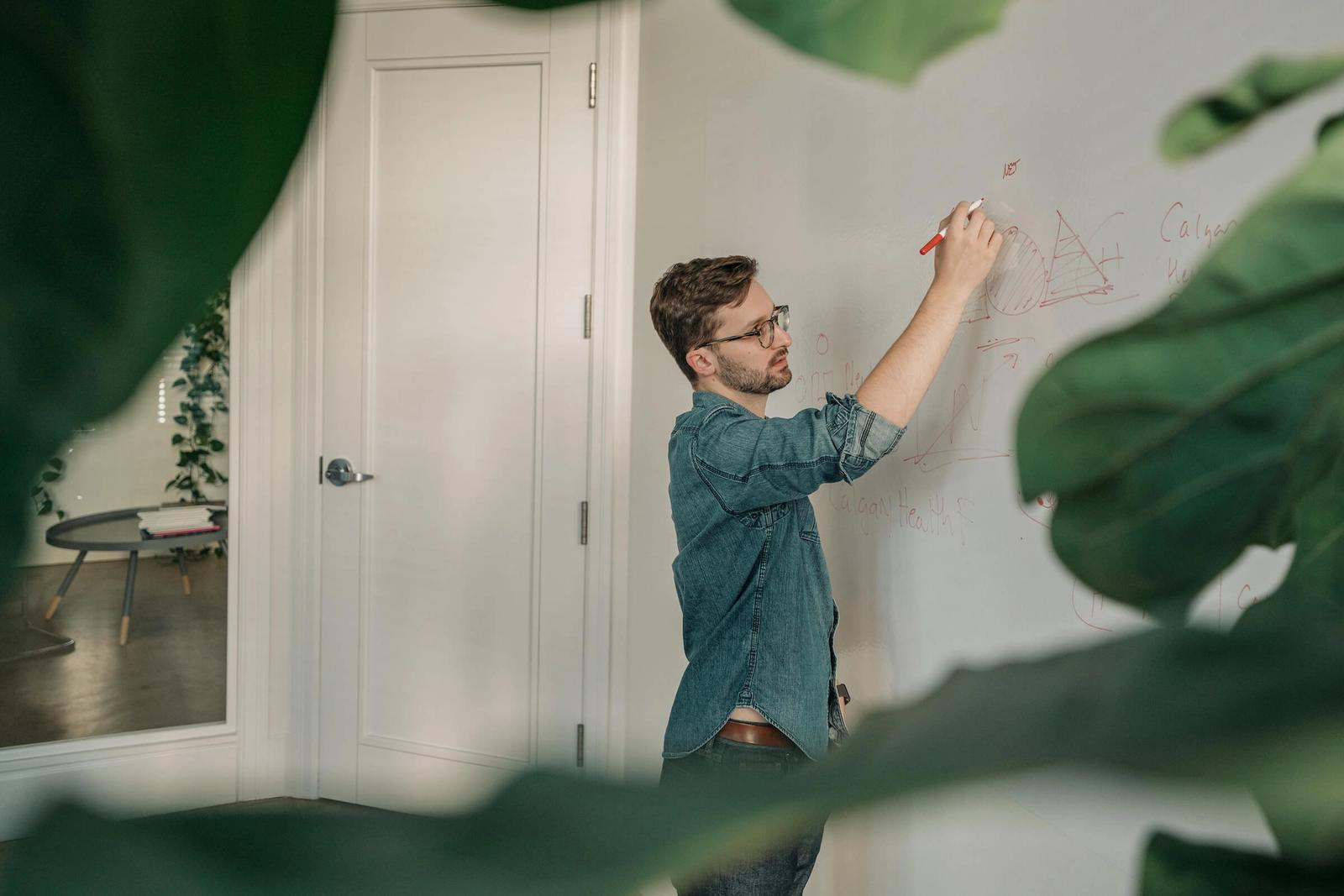 A man in a denim shirt sketching a diagram on a whiteboard, framed by leaves in the foreground.