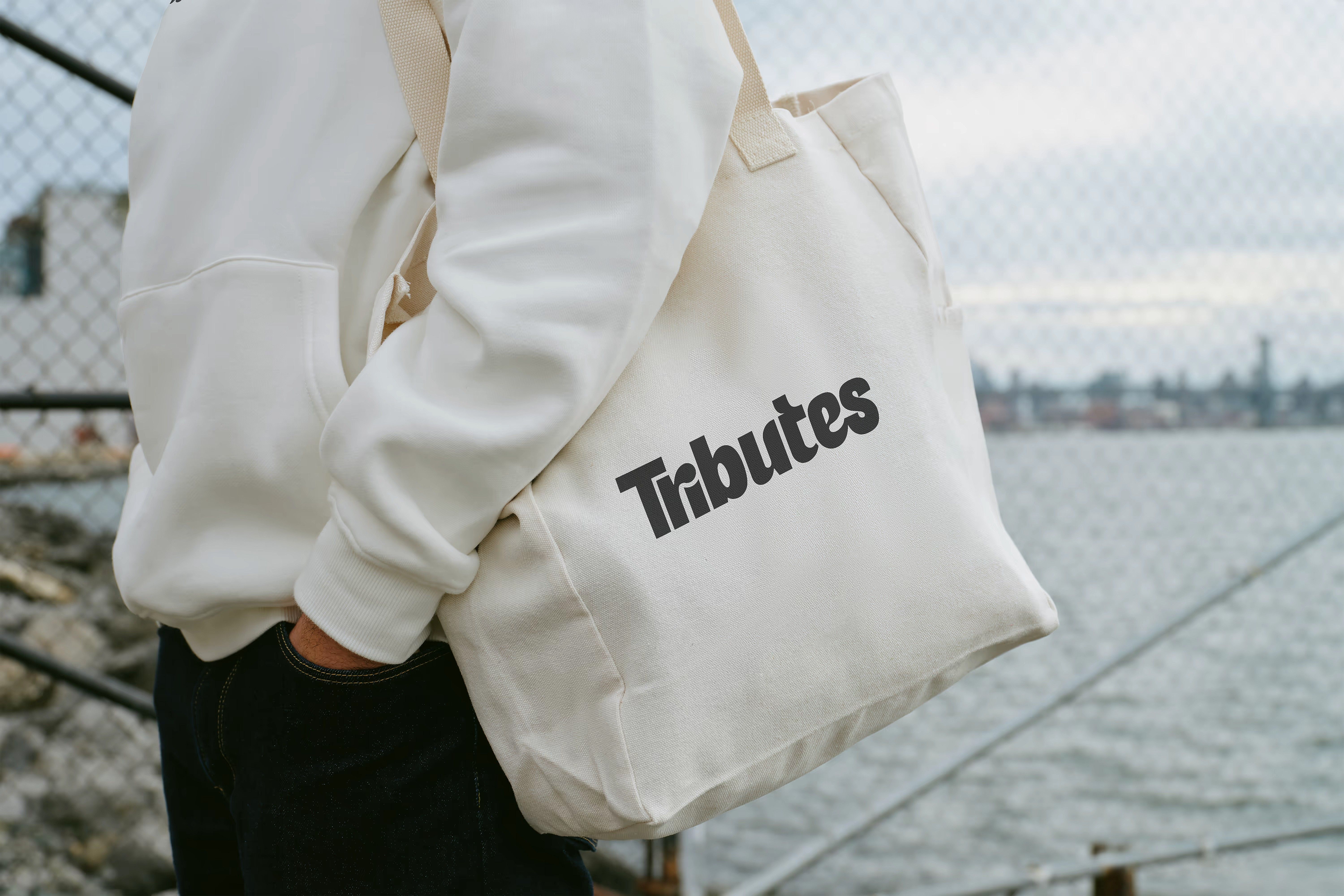 A person carrying a white canvas tote bag printed with the Tributes wordmark beside a waterfront.