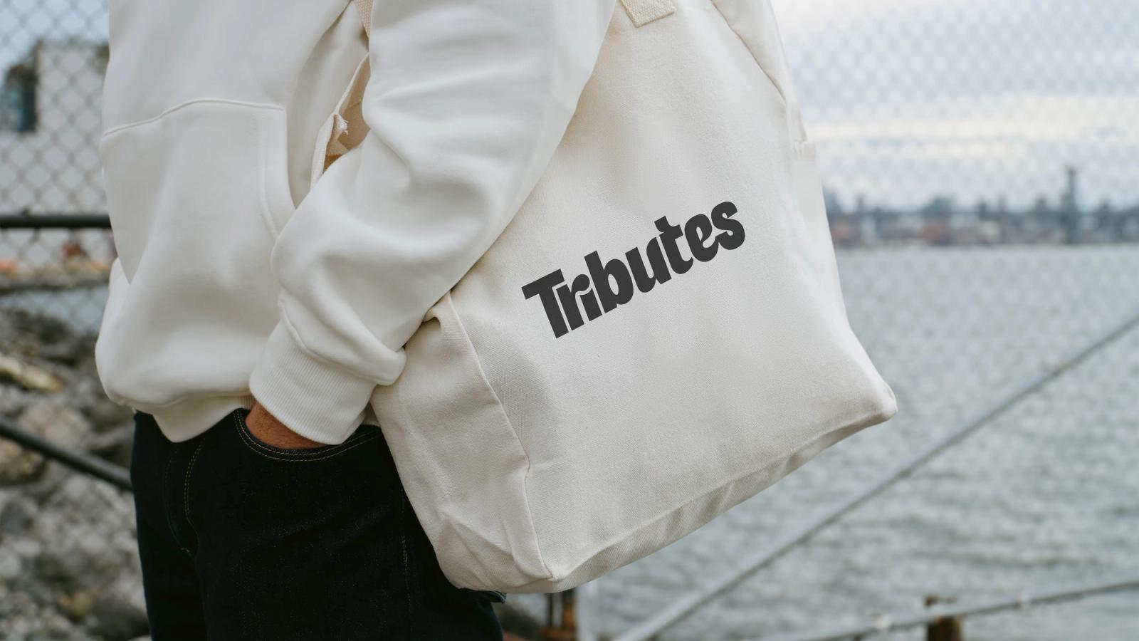 A person carrying a white canvas tote bag printed with the Tributes wordmark beside a waterfront.