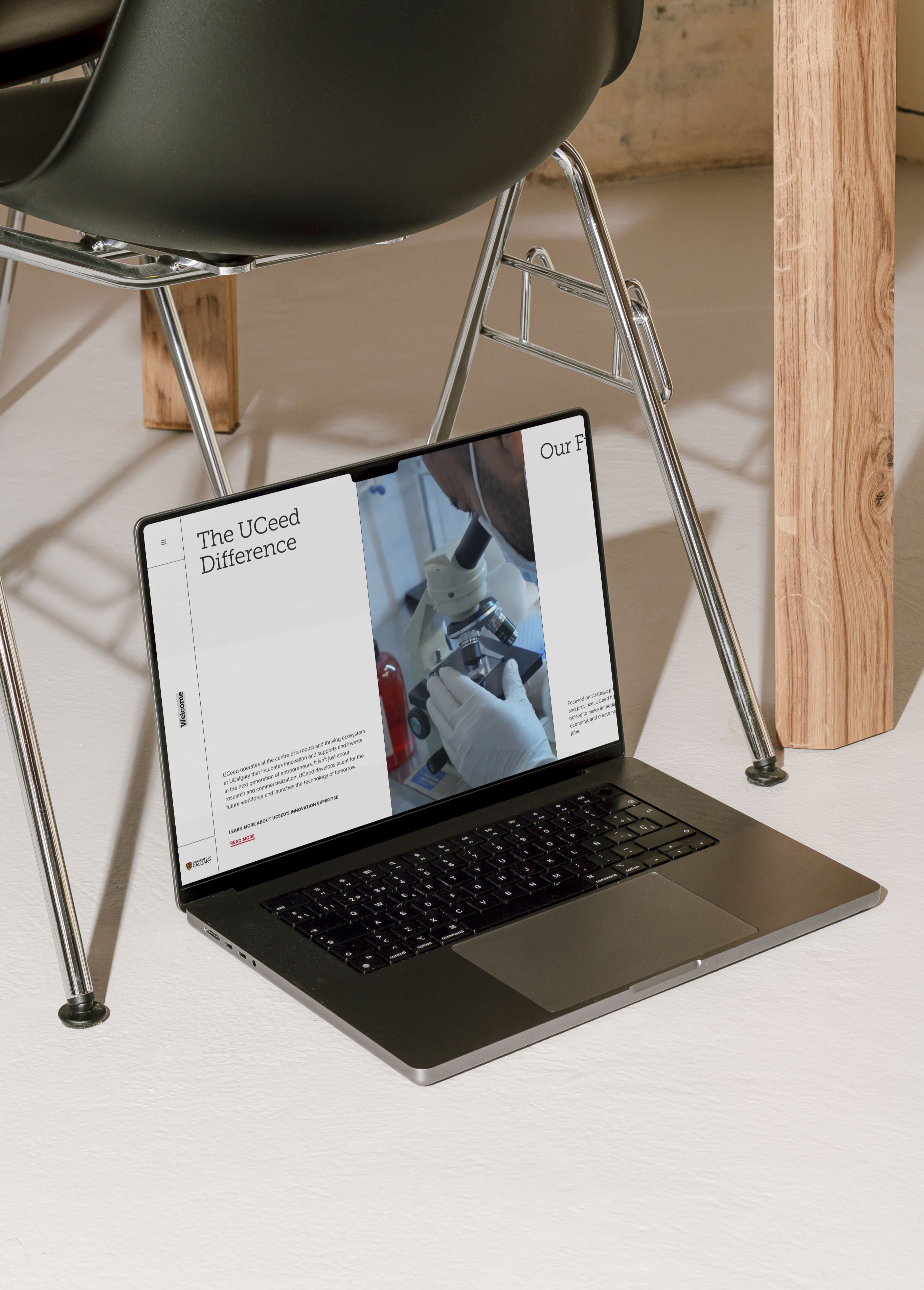 MacBook Pro resting on the floor beneath a chair, displaying the UCeed website's 'The UCeed Difference' page with a photo of a gloved researcher at a microscope.