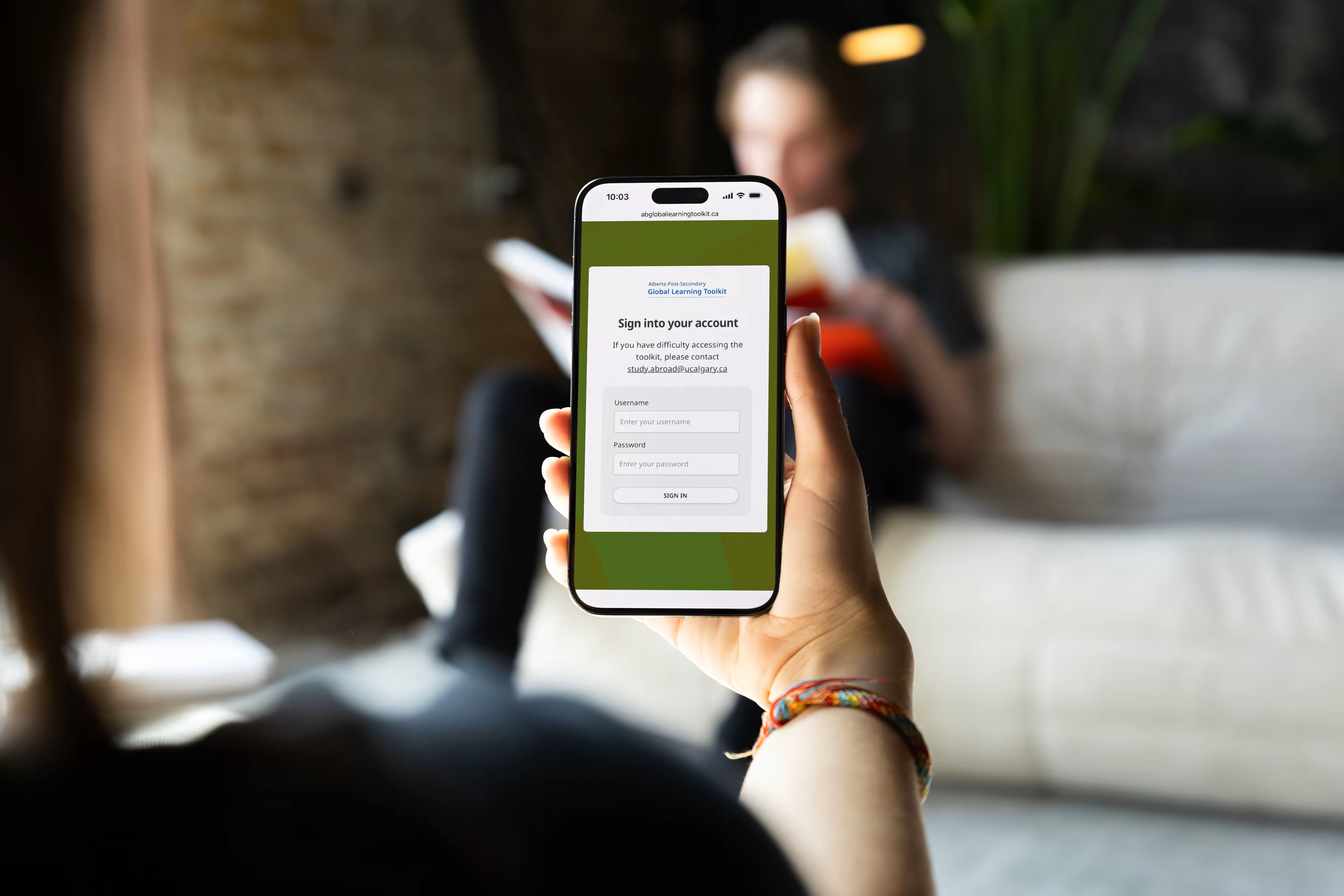 A person on a couch holding a phone that shows the sign-in screen for the Alberta Post-Secondary Global Learning Toolkit.