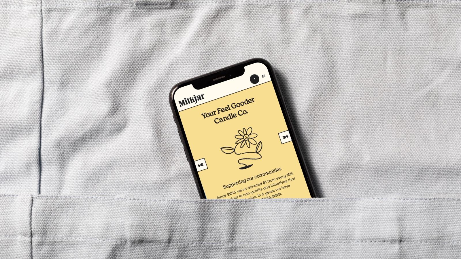 Smartphone tucked into a grey linen pocket displaying the yellow Milkjar website page reading 'Your Feel Gooder Candle Co.' with a line-drawn flower.