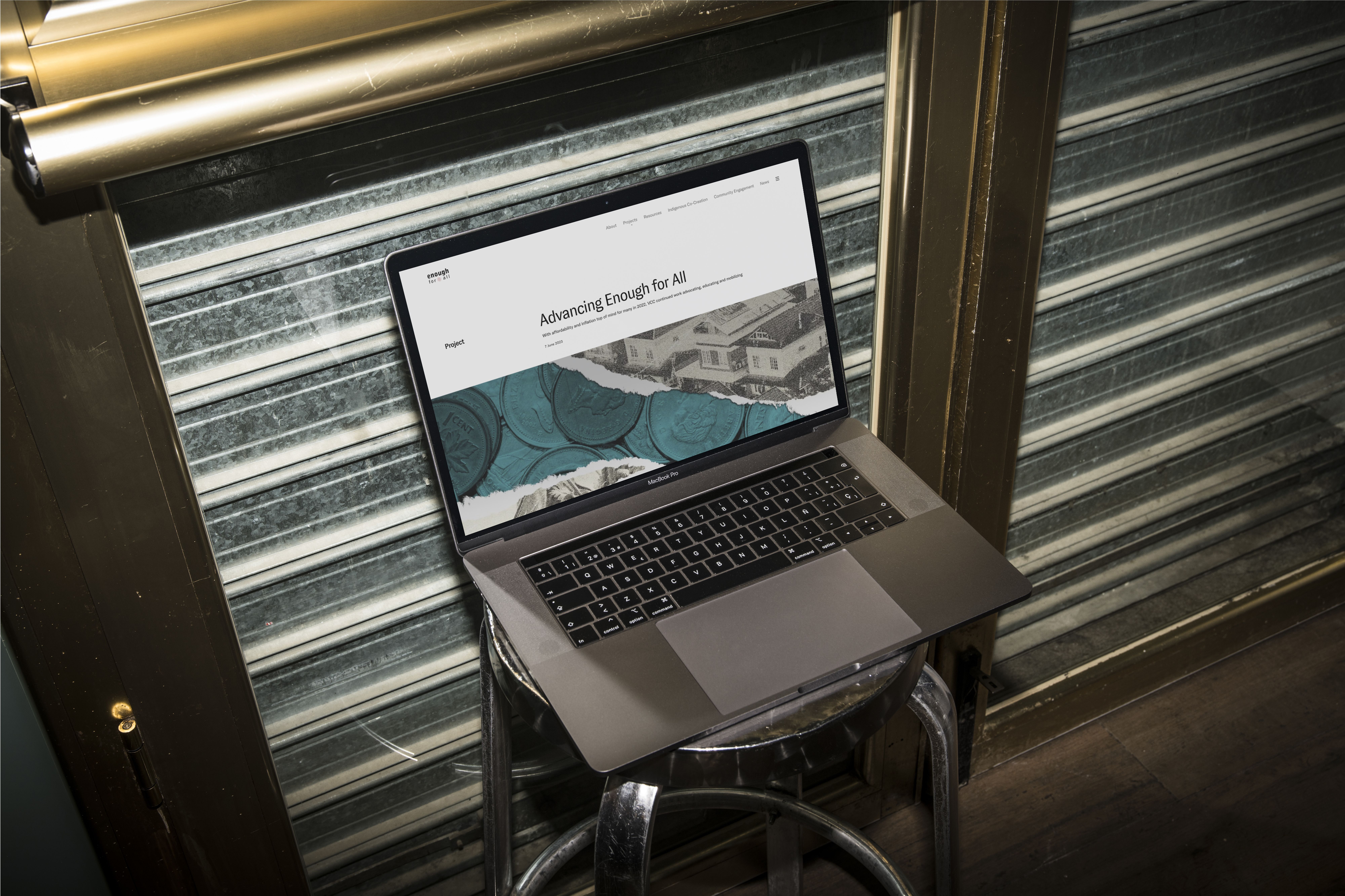 Open MacBook Pro on a metal stool against a corrugated metal shutter, displaying the 'Advancing Enough for All' webpage with a coins-and-houses collage.