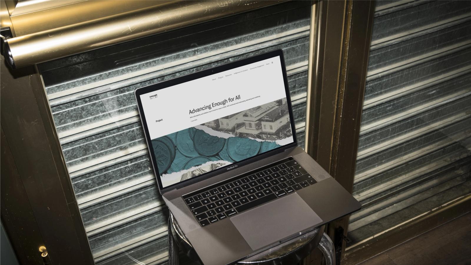 Open MacBook Pro on a metal stool against a corrugated metal shutter, displaying the 'Advancing Enough for All' webpage with a coins-and-houses collage.