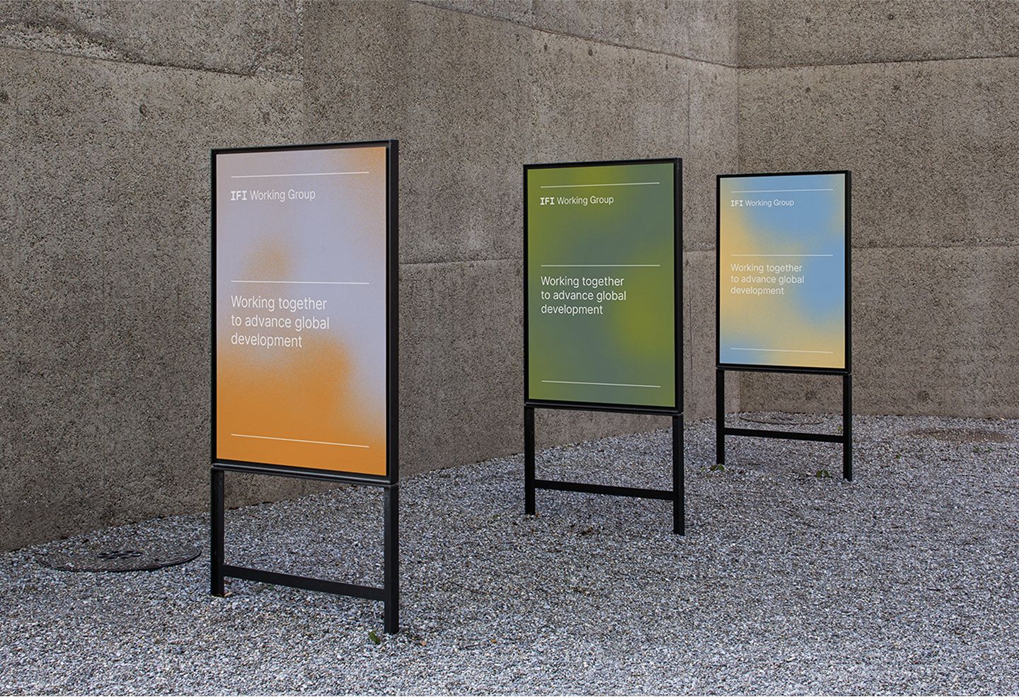 Three freestanding sidewalk poster stands on gravel against a concrete wall, each showing an 'IFI Working Group' poster reading 'Working together to advance global development.'