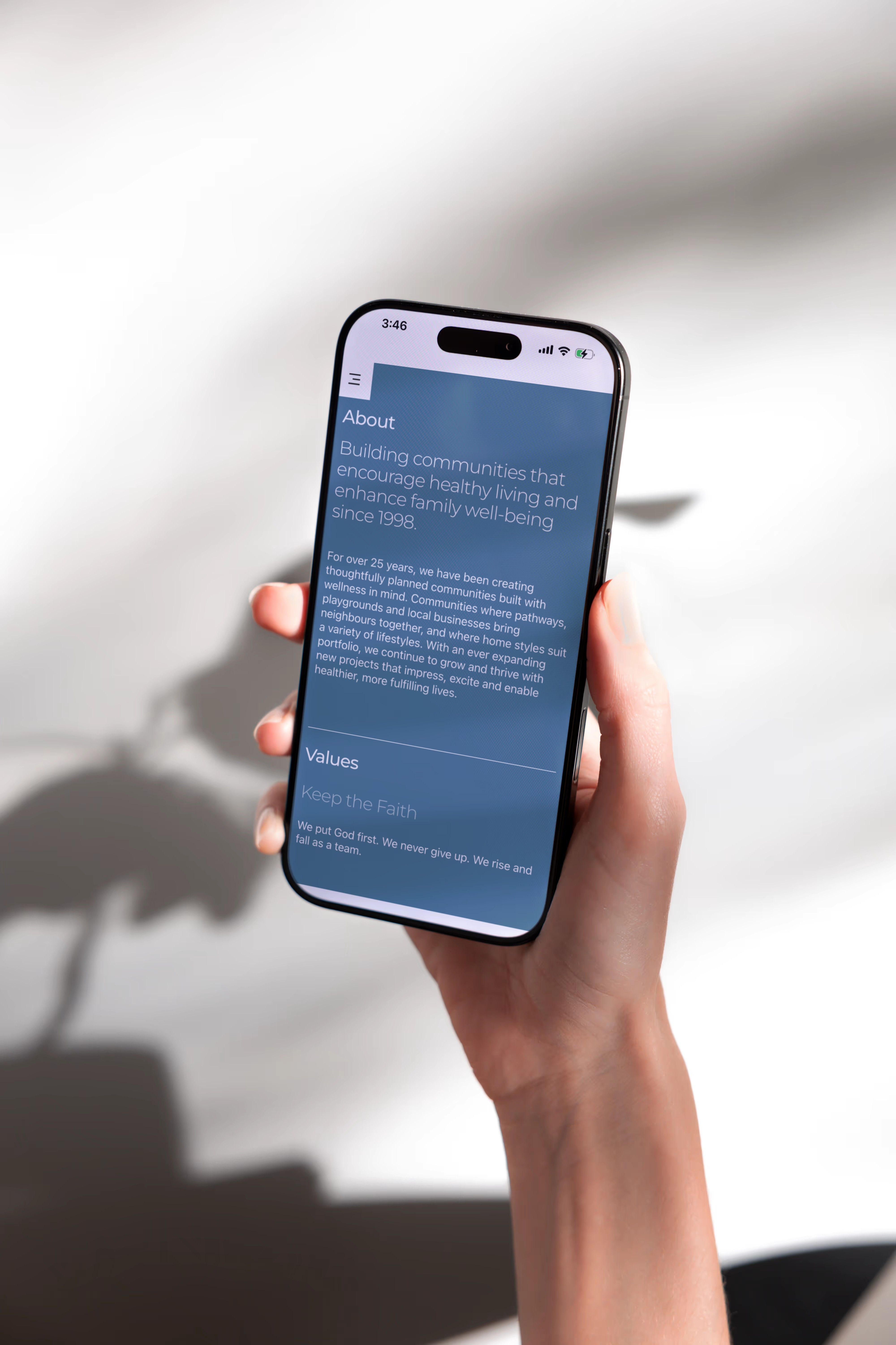 A hand holding a phone that shows the About page of a Partners Development Group microsite, in shadow against a white wall.